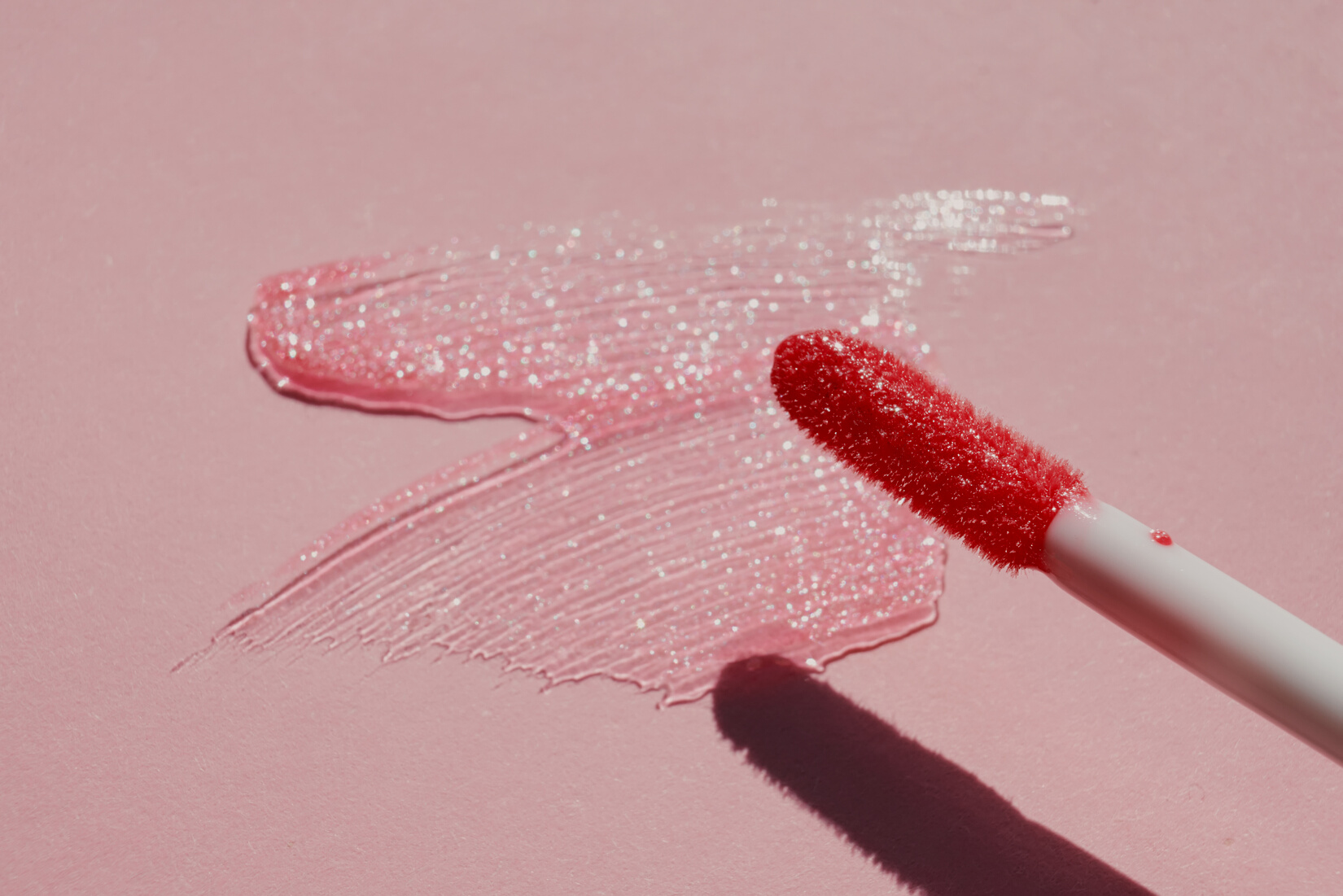 Smears of shining lip gloss and lip gloss brush on pink background, hard shadows